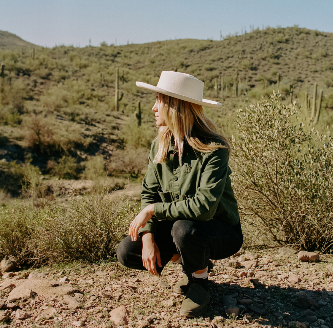 A person in a green jacket and white hat squats on rocky terrain, gazing towards cacti-dotted hills under the clear Peoria AZ sky, reminiscent of the landscapes near Saddleback.