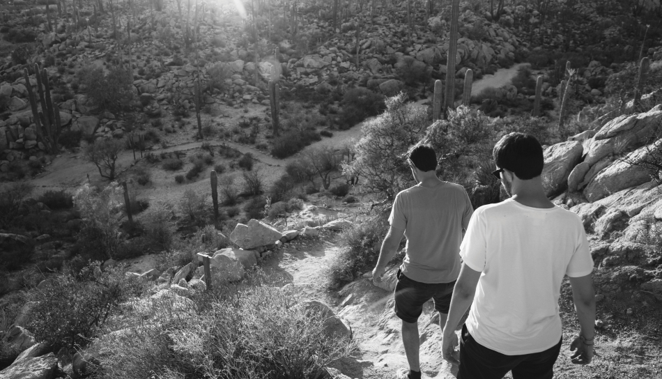 Two people walking down a rocky path in a desert landscape, Saddleback Mountain rising majestically in the background, surrounded by cacti and sunlight.